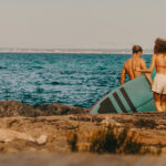 Pärchen Läuft Am Strand Richtung Meer, Frau Trägt Board Und Paddel. Pärchen Läuft Am Strand Richtung Meer, Frau Trägt Board Und Paddel.
